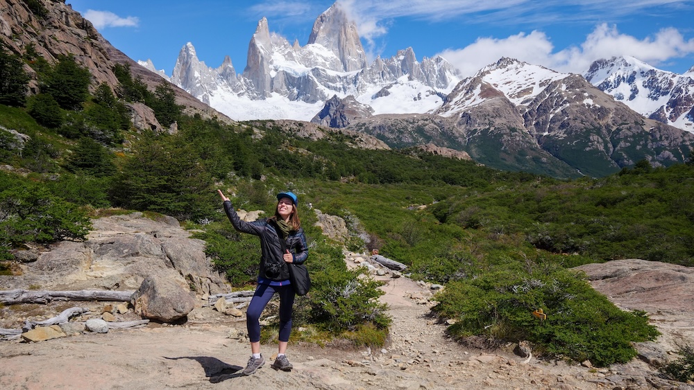 El Chaltén, Patagonia, Argentina, with Audrey Bergner standing on the hiking trail playfully presenting Mount Fitz Roy’s snow-covered granite spires rising dramatically behind her under bright blue skies
