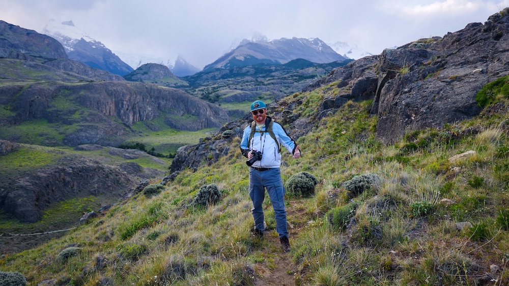 El Chaltén, Patagonia, Argentina hillside trail with Nomadic Samuel giving a thumbs-up beside rugged terrain and distant Fitz Roy peaks, showing a rewarding payoff viewpoint reached via moderate hiking conditions