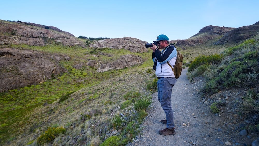 El Chaltén, Patagonia, Argentina trail scene with Nomadic Samuel stopping mid-hike to photograph rugged hills and green valleys, showing how photo breaks are part of the experience and why hiking days often take longer than planned in this dramatic landscape.