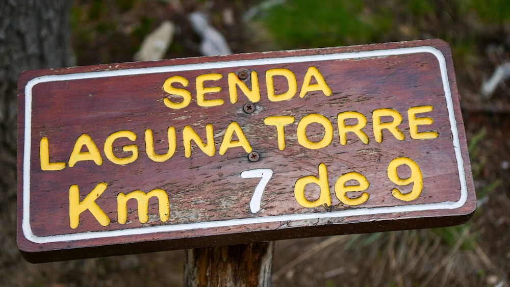 Laguna Torre kilometer marker showing pacing distance on an El Chaltén hiking trail El Chaltén, Patagonia, Argentina: a wooden trail sign reads “Senda Laguna Torre 7 de 9 km,” showing how kilometer markers help hikers track pacing, manage energy, and decide whether to continue, slow down, or turn back during long treks.