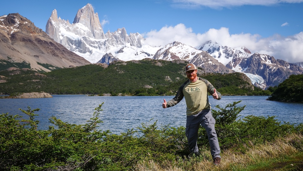 El Chaltén, Patagonia, Argentina lakeshore at Laguna Capri with a traveler giving a thumbs-up and Fitz Roy towering behind, showing the scenic payoff reached after a moderate hiking trail with steady elevation gain