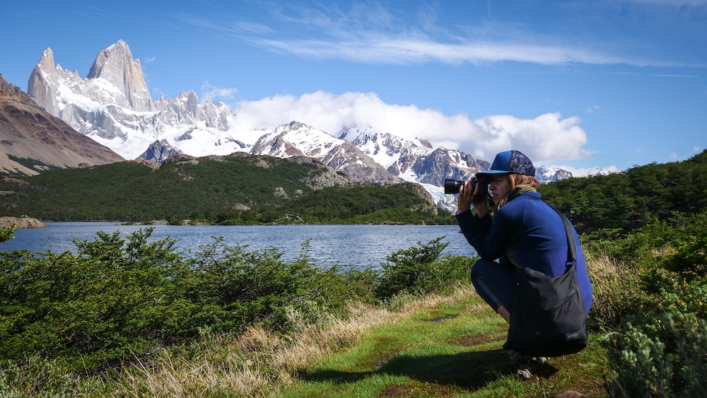 El Chaltén Patagonia Argentina Laguna Capri viewpoint with Mount Fitz Roy rising above the lake as Audrey Bergner of That Backpacker crouches with a camera capturing the dramatic alpine scenery inside Los Glaciares National Park.
