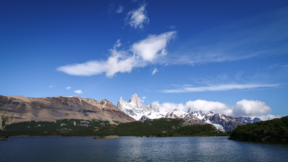 Laguna Capri in El Chaltén, Patagonia, Argentina, featuring wide-angle lake views with Mount Fitz Roy’s snow-covered granite spires rising in the distance beneath sweeping blue skies and drifting clouds
