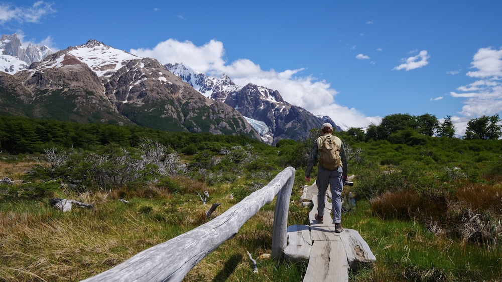 Laguna de los Tres hike in El Chaltén with Nomadic Samuel approaching Fitz Roy El Chaltén, Patagonia, Argentina — Nomadic Samuel hikes toward Laguna de los Tres along a rustic wooden boardwalk with Fitz Roy towering ahead, capturing the epic scale of the trail and the growing hunger before a well-earned post-hike meal.