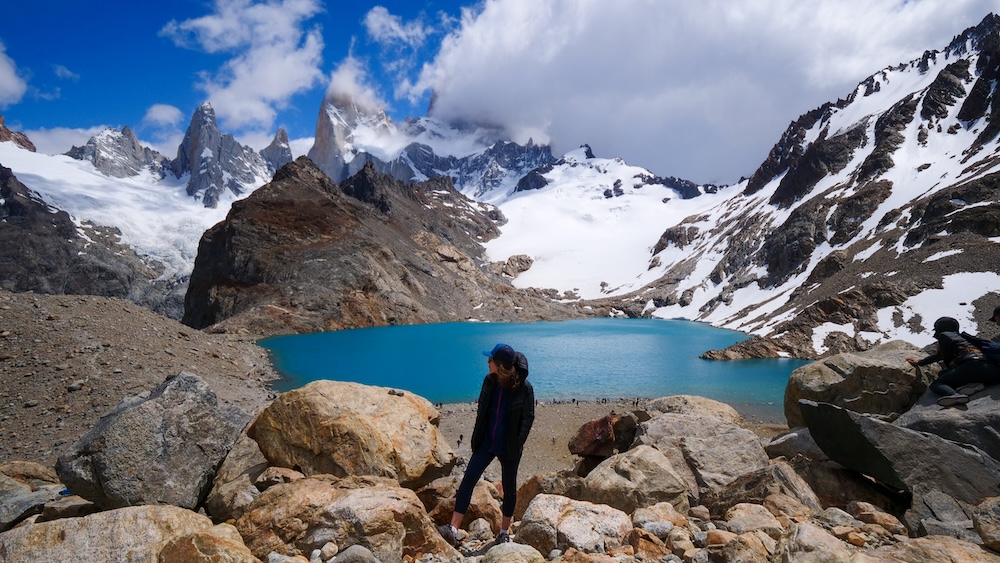 Audrey Bergner bundled in layers at Laguna de los Tres beneath Mount Fitz Roy in El Chaltén El Chaltén, Patagonia, Argentina: Audrey Bergner bundled in warm layers at Laguna de los Tres, standing beside the turquoise glacial lake beneath Mount Fitz Roy, showing how cold and windy conditions can feel even on clear hiking days.