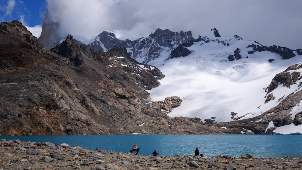 El Chaltén, Patagonia, Argentina turquoise lake at Laguna de los Tres with hikers resting on the rocky shore beneath steep snow-covered peaks, illustrating the dramatic payoff reached after a long and physically demanding hiking trail