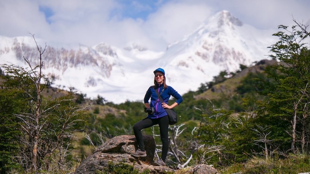El Chaltén, Patagonia, Argentina hiking scene at Laguna Torre, with Audrey Bergner standing on a rock posing confidently as snowcapped Fitz Roy peaks rise behind, showcasing her Argentina travel expertise.