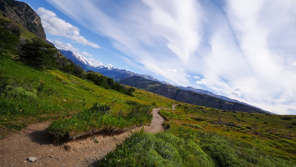 A quiet stretch of the Laguna Torre trail in El Chaltén, Patagonia, Argentina, with a winding dirt path through green hills, distant snowcapped peaks, and dramatic clouds, capturing the rare feeling of hiking completely alone in a famously popular destination.