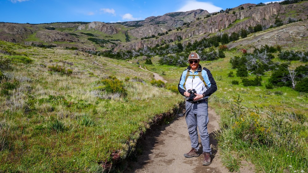 Laguna Torre trail hiking with Nomadic Samuel in El Chaltén El Chaltén, Patagonia, Argentina — Nomadic Samuel hikes the Laguna Torre trail with camera in hand, trekking through green valleys and rocky cliffs while documenting one of Patagonia’s most iconic mountain routes.