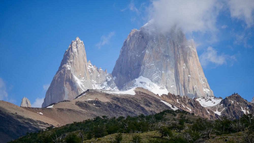 El Chaltén, Patagonia iconic Mount Fitz Roy rising above forested hills, with sheer granite spires, lingering snow, and drifting clouds, capturing the dramatic scale and raw beauty that define Argentina’s most famous hiking landscape.