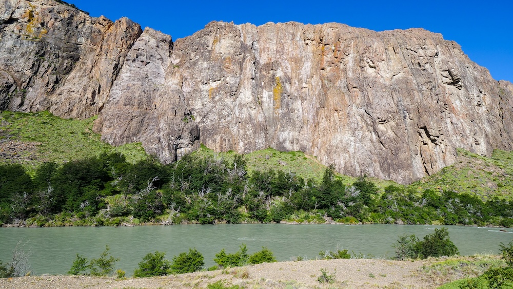 El Chaltén, Patagonia, Argentina: a peaceful river flows beneath towering rock cliffs and green forest, a gentle rest-day riverside walk that still delivers classic Patagonian scenery without a long hike.