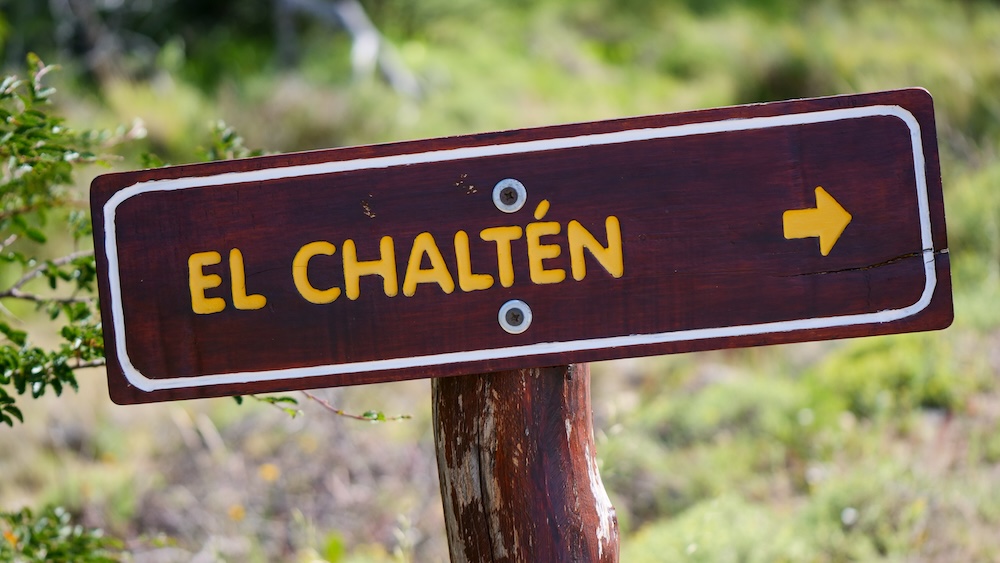 El Chaltén wooden sign – hiking capital of Patagonia and base for epic day trips El Chaltén, Patagonia, Argentina wooden trail sign pointing toward town, set against green hillside scenery in soft daylight—an iconic marker welcoming travelers to the hiking capital of Patagonia and the perfect base for day trips.