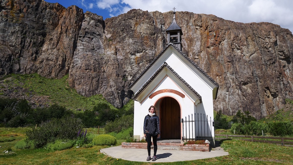 El Chaltén, Patagonia, Argentina: Audrey Bergner stands in front of the Capilla de los Escaladores chapel beneath dramatic cliffs, enjoying a peaceful town-wander rest day surrounded by mountain scenery.