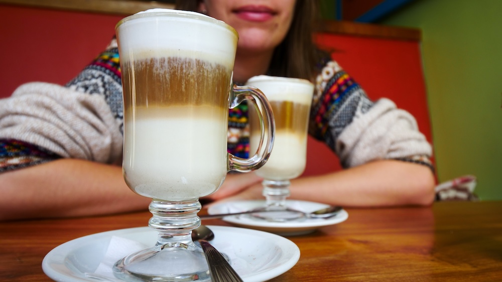 El Chaltén, Patagonia — layered lattes on a wooden café table with Audrey Bergner resting behind them, enjoying a warm drink on a weather day, capturing the cozy café culture that makes rest days in Argentina’s hiking capital so satisfying.