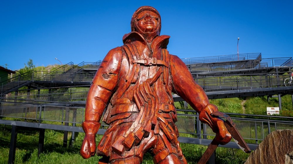 El Chaltén, Patagonia: wooden hiker monument statue depicting a backpacked trekker with ice axe, celebrating the town’s hiking culture and reputation as Argentina’s trekking capital under clear blue skies.
