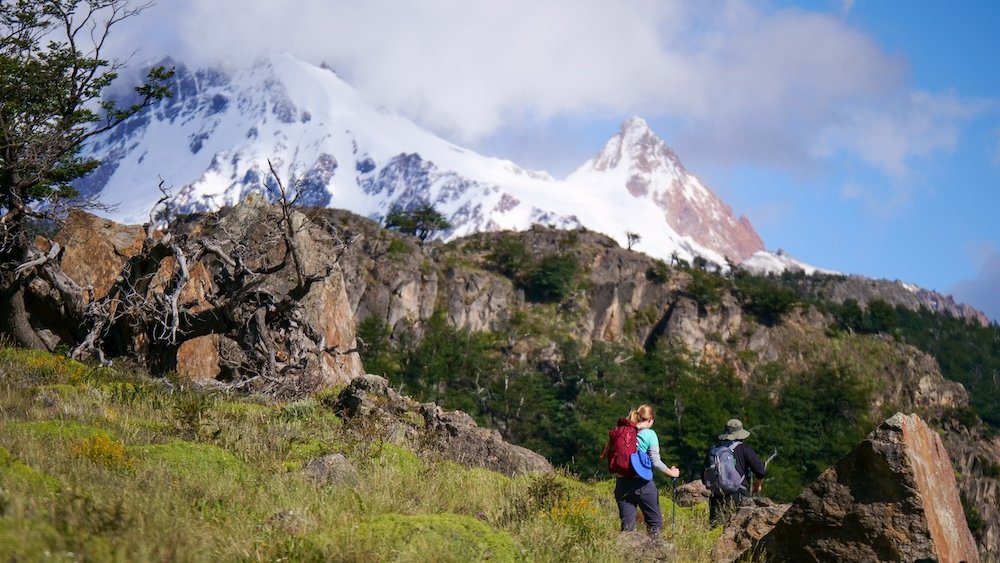 Hiking Laguna de los Tres in El Chaltén, Patagonia, with trekkers walking through alpine terrain toward Mount Fitz Roy, featuring rocky hillsides, lenga forest, snow-covered peaks, and clear mountain weather along one of Patagonia’s most iconic trails.