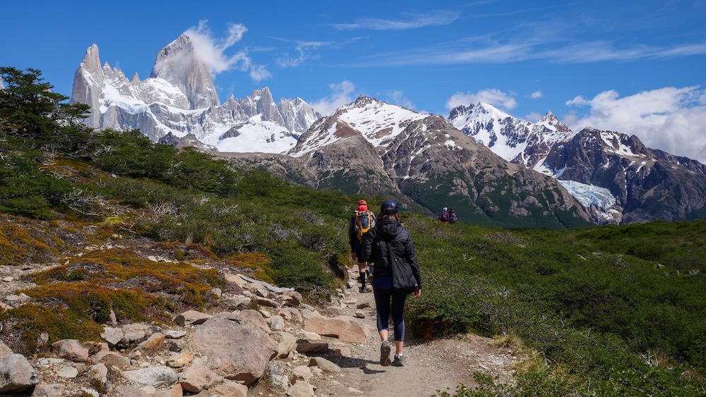 El Chaltén, Patagonia — hikers walk the rocky trail toward Mount Fitz Roy, capturing the classic Laguna de los Tres route through low shrubs and mountain scenery in Los Glaciares National Park, Argentina.