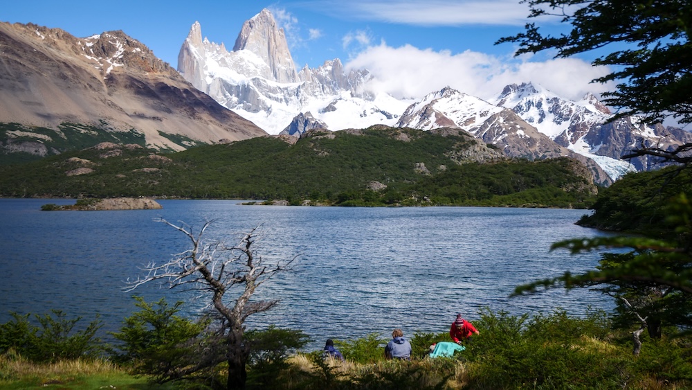 El Chaltén Patagonia Argentina view of Laguna Capri with hikers resting on the shoreline, deep blue lake water, forested hills, and the dramatic Fitz Roy massif rising in the background on a clear hiking day