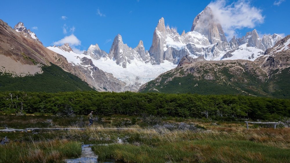 El Chaltén, Patagonia: Fitz Roy dominates the skyline above lenga forest and wetlands on the Laguna de los Tres hike, with Nomadic Samuel appearing as a tiny hiker on the trail, emphasizing the dramatic scale of the mountains.
