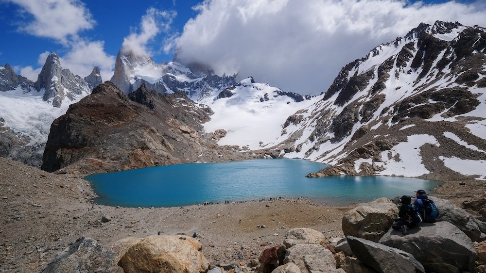 Laguna de los Tres payoff viewpoint in El Chaltén, Patagonia, showing a turquoise glacial lake beneath Mount Fitz Roy with snow-covered peaks, hikers resting on rocks, and dramatic alpine scenery after completing the steep final climb of this iconic boss-level hike.