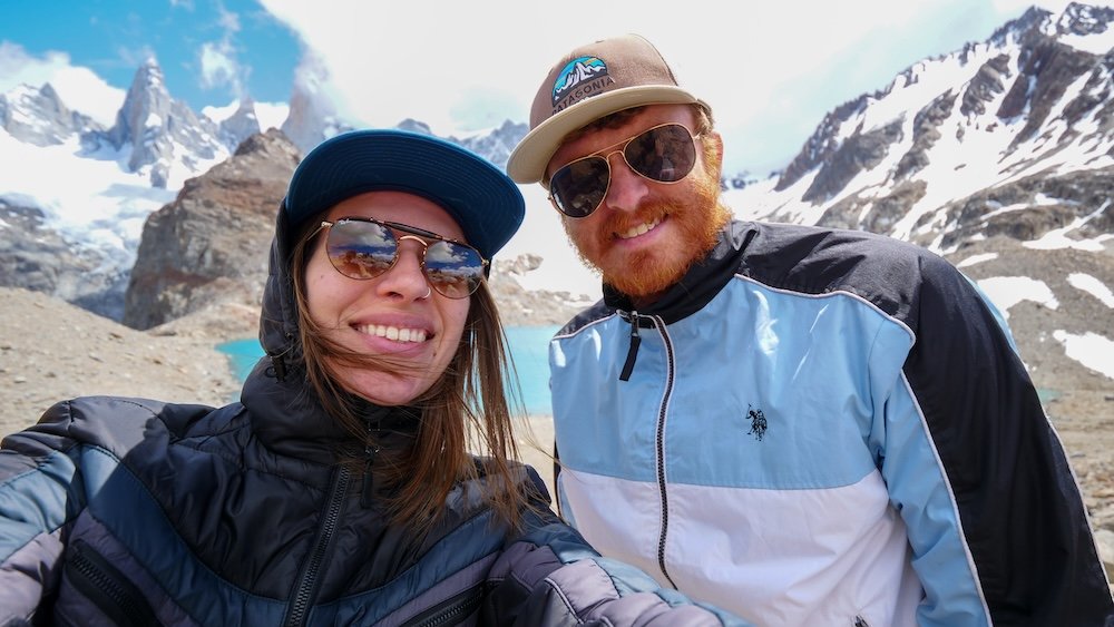 Laguna de los Tres in El Chaltén, Patagonia, featuring Nomadic Samuel and Audrey Bergner smiling at the iconic Fitz Roy payoff viewpoint, with turquoise glacial lake, jagged granite peaks, snow-covered mountains, and strong Patagonian wind on a clear hiking day.