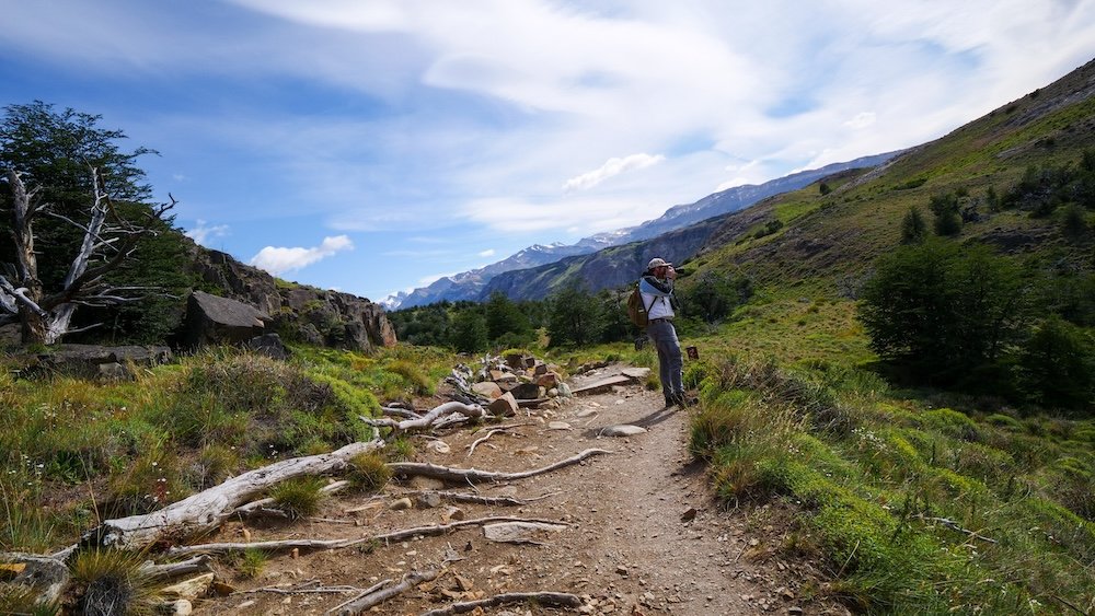 Nomadic Samuel stopping to take photos while hiking the Laguna Torre trail in El Chaltén, Patagonia, surrounded by rolling green hills, rocky trail, alpine vegetation, and wide valley views during a scenic break on one of Patagonia’s most enjoyable day hikes.