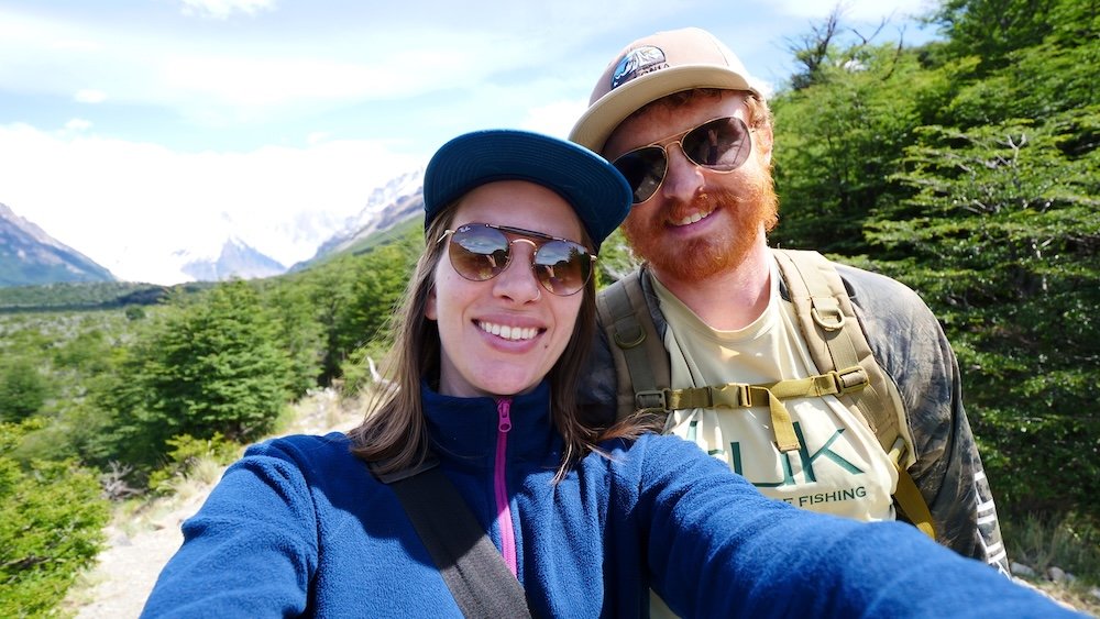 Nomadic Samuel and Audrey Bergner smiling while hiking the Laguna Torre trail in El Chaltén, Patagonia, with green lenga forest, backpacks, and distant mountain peaks behind them on a clear trekking day in Los Glaciares National Park, Argentina.