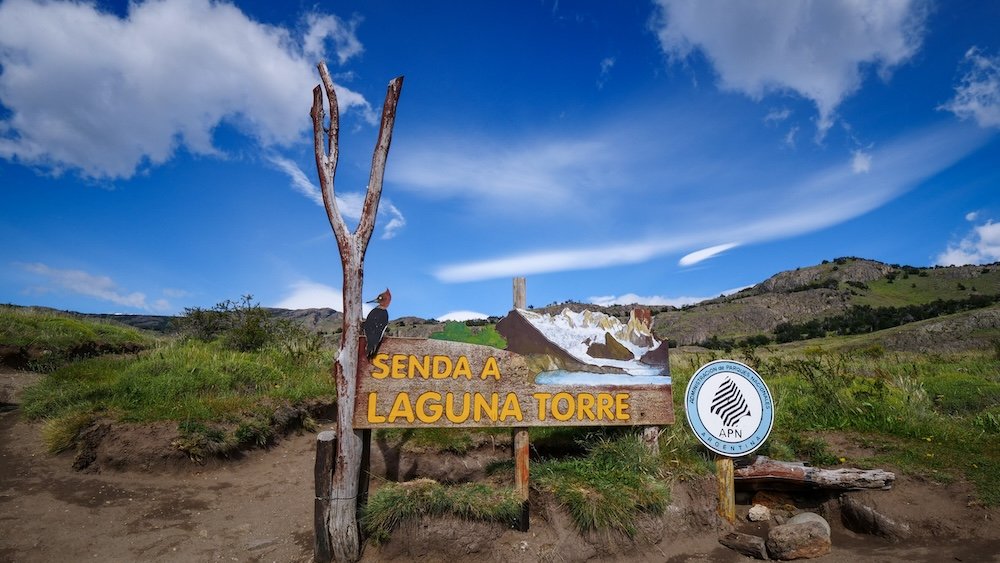 El Chaltén, Patagonia: the wooden “Senda a Laguna Torre” trailhead welcome sign marks the start of the Laguna Torre hike, surrounded by open hills, dirt paths, and blue skies on a calm trekking day.
