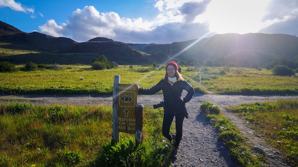 El Chaltén, Patagonia, Argentina: a smiling That Backpacker Audrey Bergner traveler stands beside the Miradores trail sign at the Mirador de los Cóndores trailhead, ready for an easy sunset hike with valley views and glowing mountain scenery.