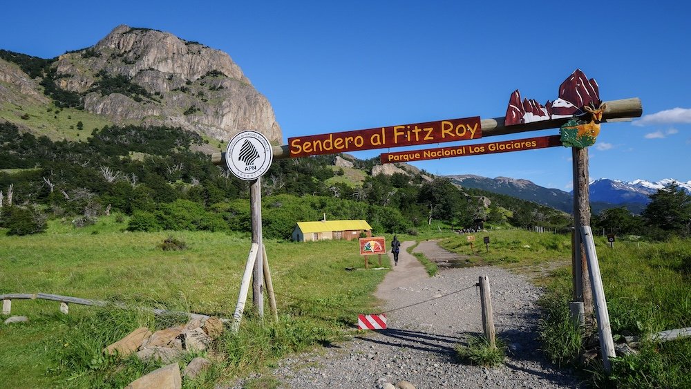 El Chaltén, Patagonia: the Sendero al Fitz Roy trailhead sign marks the main portal hike for Laguna de los Tres, with a gravel path leading into green meadows, forested slopes, and rugged mountains on a clear day.
