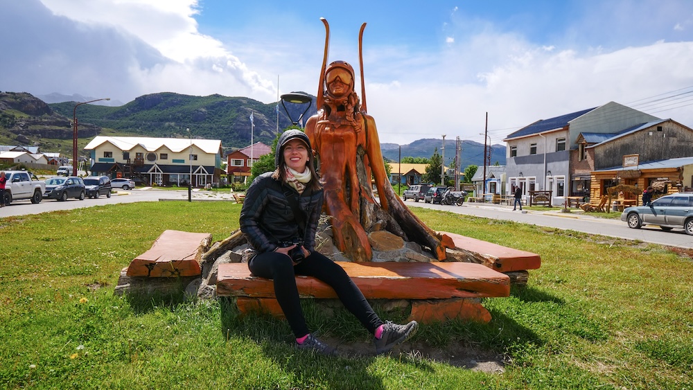 El Chaltén town center in Patagonia with Audrey Bergner sitting beside a carved wooden hiker sculpture, wearing layered trekking clothes and leggings, surrounded by grassy streets, colorful buildings, and mountain views in Los Glaciares National Park.