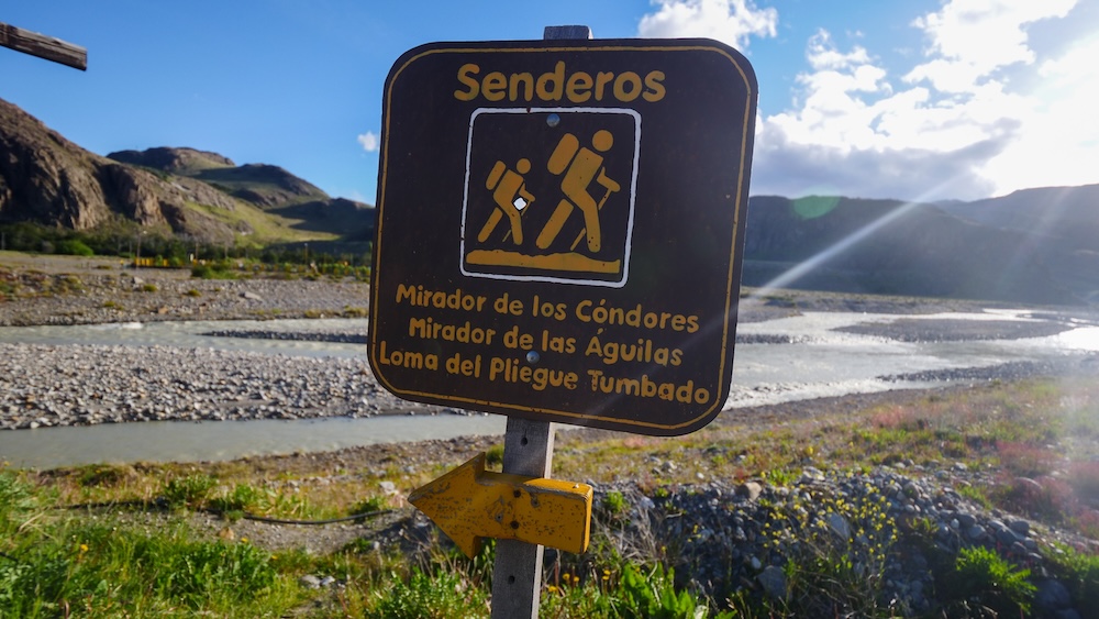 El Chaltén Patagonia hiking trail sign pointing to Mirador de los Cóndores, Mirador de las Águilas, and Loma del Pliegue Tumbado beside the Río de las Vueltas in Los Glaciares National Park near the trekking town trailheads.
