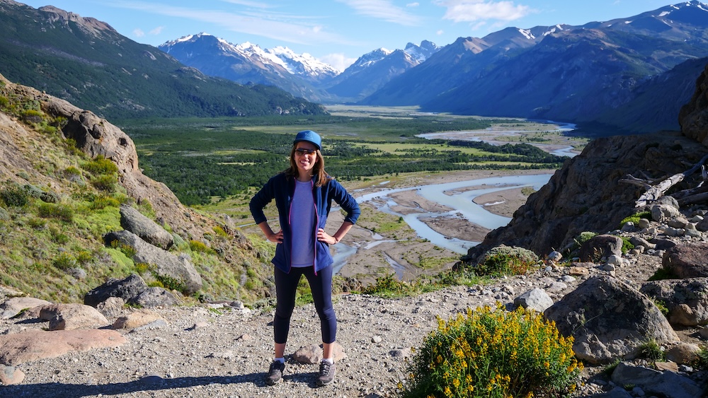 El Chaltén Patagonia valley viewpoint with Audrey Bergner standing on a rocky trail in leggings and layered hiking clothes, overlooking a winding river and vast green plains beneath snow-capped peaks in Los Glaciares National Park.