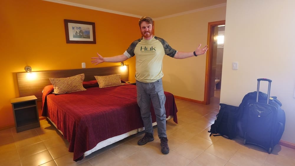 Samuel Jeffery stands with arms outstretched in a cozy orange-walled hotel room in El Chaltén, Santa Cruz, Argentina, with luggage stacked by the door and a framed mountain landscape print above the bed.