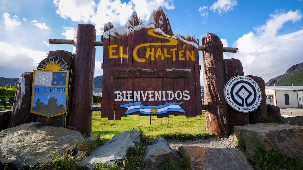 A wooden welcome sign painted with mountain peaks reads "El Chaltén, Capital Nacional del Trekking, Bienvenidos" beneath a blue sky at the entrance to El Chaltén, Santa Cruz, Argentina.
