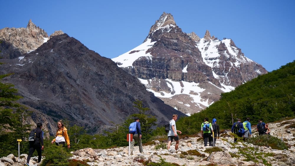 Hikers on a Rocky Trail Below Glacial Peaks in El Chaltén, Santa Cruz, Patagonia, Argentina Hikers walk a rocky trail beneath sharp snow-streaked peaks in El Chaltén, Santa Cruz, with low forest and rugged mountain scenery showing the trekking terrain of Patagonia, Argentina.