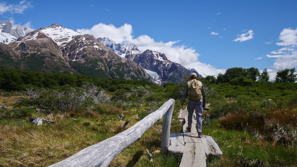 Samuel Jeffery Hiking Wooden Trail Toward Fitz Roy Glacier, El Chaltén, Santa Cruz, Patagonia Samuel Jeffery walks a wooden boardwalk trail through grassland scrub toward snow-capped Fitz Roy peaks and a hanging glacier near El Chaltén, Santa Cruz Province, in Argentine Patagonia, under bright blue sky.