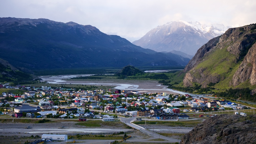 El Chaltén town seen from a high viewpoint in Patagonia, Argentina, with colorful buildings spread across the Río de las Vueltas valley beneath dramatic mountains and cloudy skies in Los Glaciares National Park.