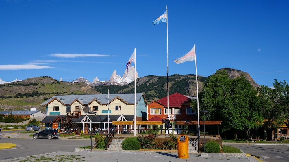 El Chaltén town center with Argentine flags and Mount Fitz Roy in the background El Chaltén town views in Patagonia, Argentina, with colorful buildings, Argentine flags waving in the foreground, and the jagged granite spires of Mount Fitz Roy rising dramatically behind the village on a clear blue-sky day.