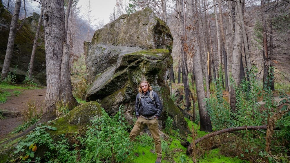 Glacial Boulders and Charred Forest Near El Hoyo, Chubut, Patagonia, with Samuel Jeffery Samuel Jeffery stands beside a massive moss-covered glacial boulder in a charred forest near El Hoyo, Chubut, Patagonia, Argentina, surrounded by blackened trunks and fresh green undergrowth.