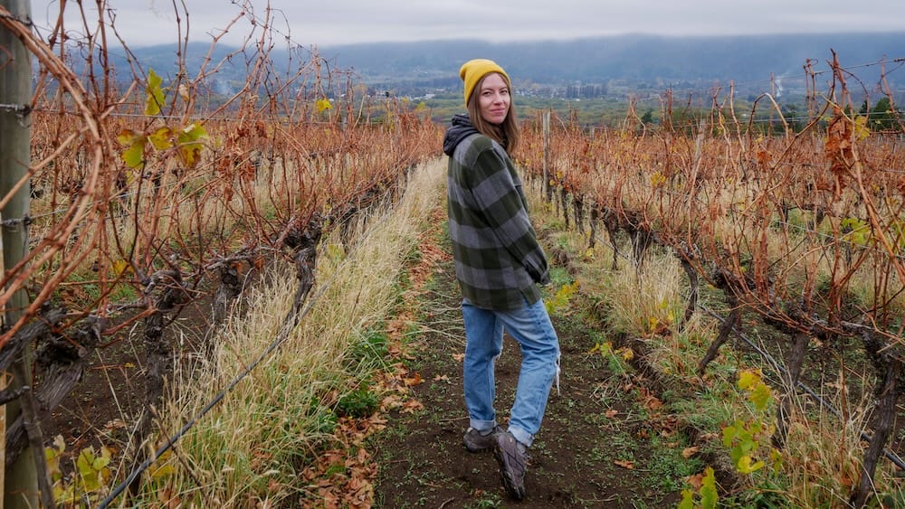 Audrey Bergner among vineyard rows in El Hoyo, Chubut, Patagonia, Argentina, with a misty valley backdrop Audrey Bergner stands between vineyard rows in El Hoyo, Chubut, Patagonia, Argentina, wearing a yellow beanie and jacket, with bare vines, earthy paths and a misty valley landscape stretching out beneath an overcast sky.