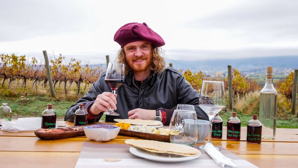 El Hoyo vineyard wine tasting in Chubut, Patagonia, Argentina, with Samuel Jeffery and an autumn picnic Samuel Jeffery raises a glass of red wine during a vineyard picnic in El Hoyo, Chubut, Patagonia, Argentina, with tasting bottles, cheeses, tableware and autumn vines beneath an overcast sky.