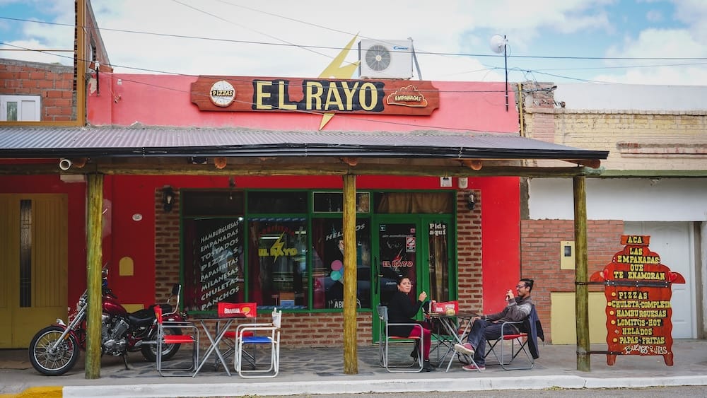 Two people take an afternoon meal break outside El Rayo, a bright red local eatery in Dolavon, Chubut, Patagonia, Argentina, with motorcycles, outdoor tables, and handwritten food signs along the storefront.