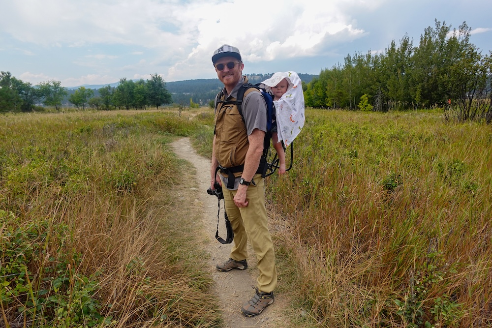Elizabeth Lake Bird Sanctuary in Cranbrook, BC during a family-friendly hike, showing Nomadic Samuel carrying baby Aurelia in a backpack carrier while walking a narrow trail through tall grasses, highlighting peaceful wetlands, open skies, and relaxed outdoor travel with a baby.
