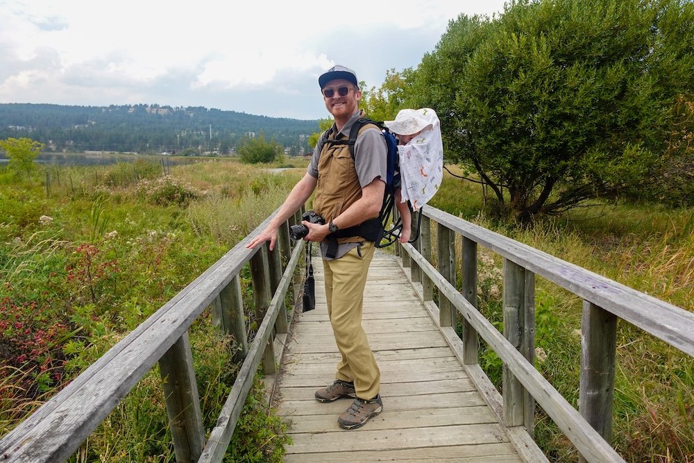 Nomadic Samuel hiking with baby Aurelia at Elizabeth Lake in Cranbrook Nomadic Samuel Jeffery stands on a wooden boardwalk overlooking the wetlands of Elizabeth Lake in Cranbrook, BC, carrying baby Aurelia in a hiking backpack while taking in the early September scenery under soft mountain light.