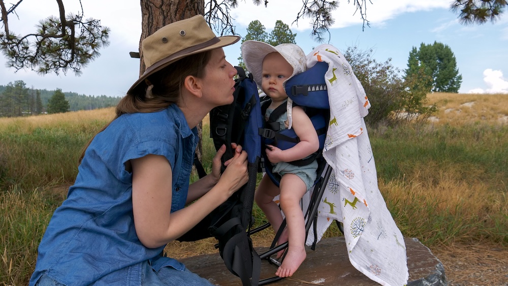 Elizabeth Lake in Cranbrook, BC during a relaxed family hike, showing That Backpacker Audrey Bergner pausing beside the trail while baby Aurelia sits comfortably in a backpack carrier, surrounded by grassy wetlands and pine trees that highlight how peaceful and baby-friendly this nature walk feels.
