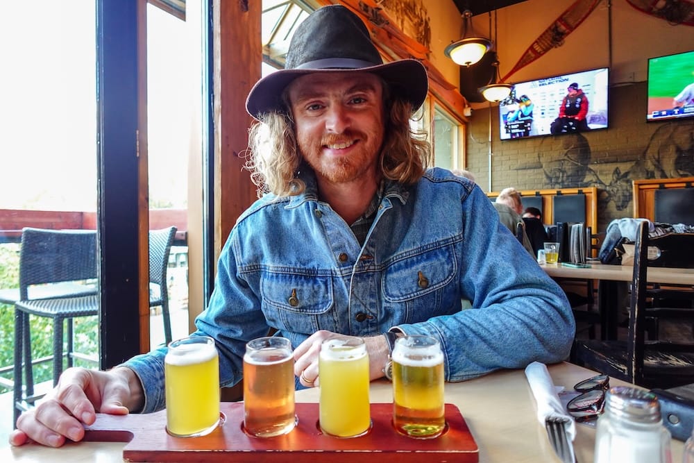 Samuel Jeffery with a Beer Flight at Elk & Oarsman in Banff, Alberta, Canada Samuel Jeffery seated at Elk & Oarsman in Banff, Alberta, Canada, with a wooden tasting flight of four small beers on the table and the casual pub interior visible behind him.