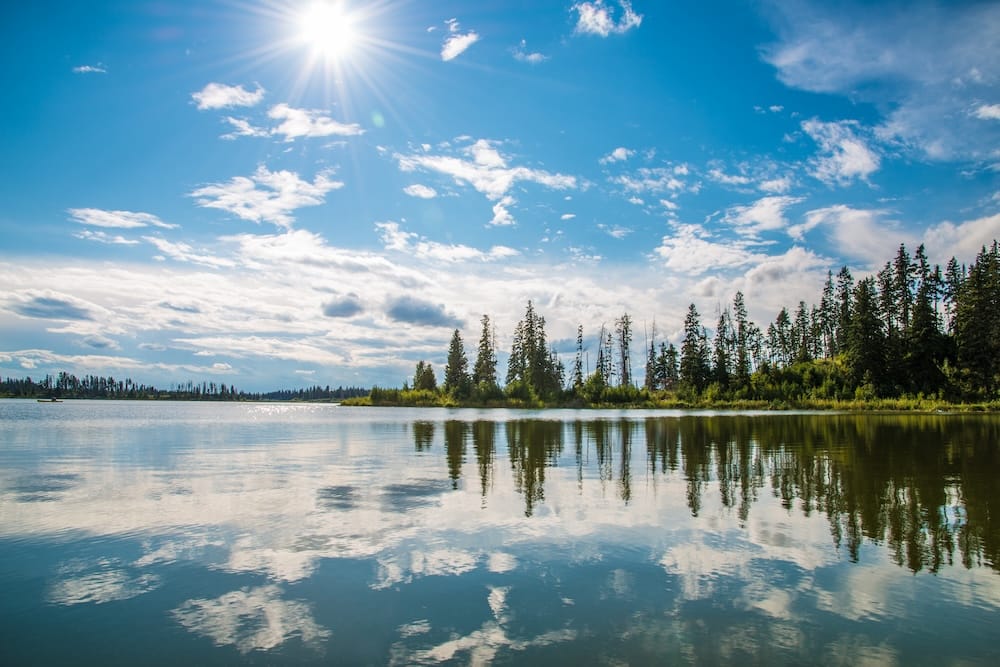 Calm lake scenery in Elk Island National Park, Alberta, Canada, with evergreen forest along the shoreline, reflections on the water and bright sun breaking through a blue sky.
