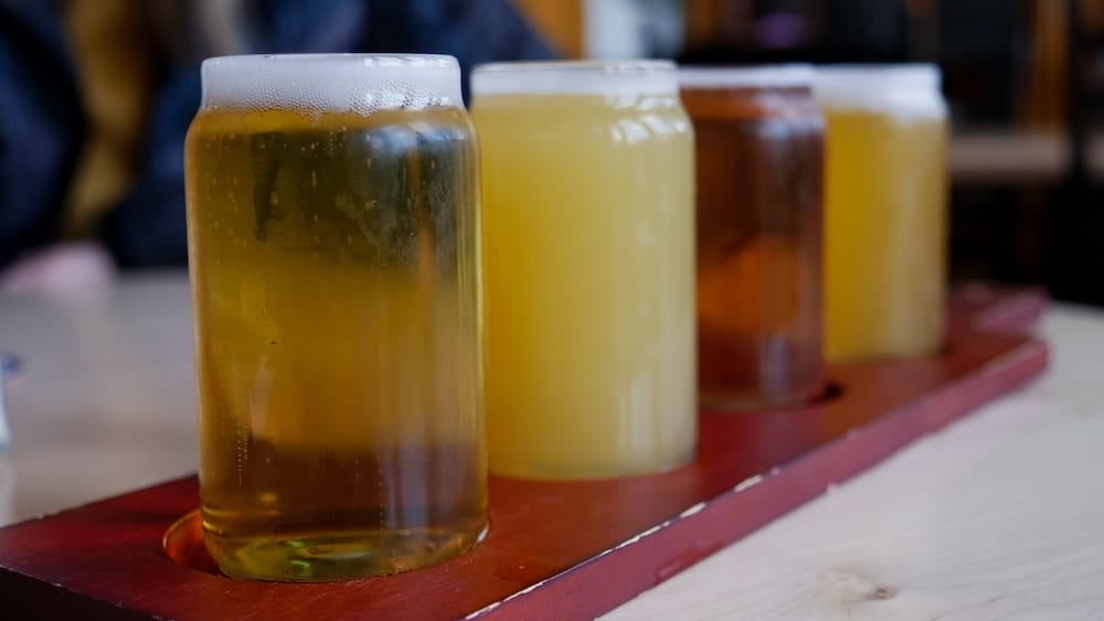 Close-up of a four-glass beer flight on a wooden tasting paddle at Elk & Oarsman in Banff, Alberta, Canada, showing pale and amber beers on a pub table with the restaurant interior softly blurred in the background.