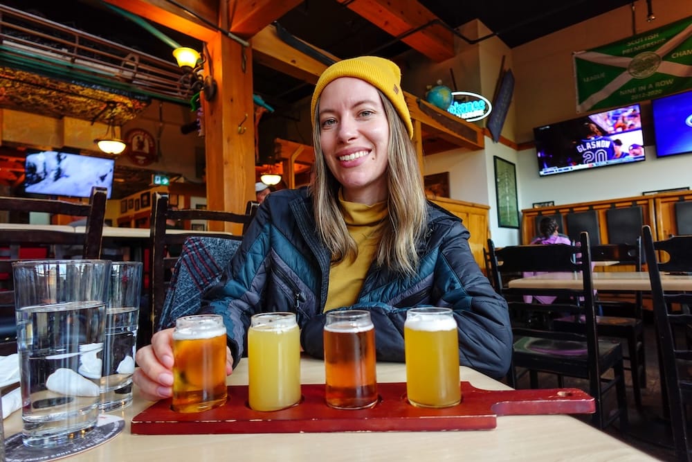 Audrey Bergner sits inside Elk & Oarsman in Banff, Alberta, Canada, holding one glass from a four-beer flight on the table, with timber interiors, pub seating and sports screens visible behind her.